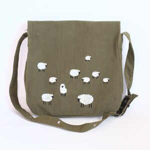 May include: Olive green messenger bag with a flap closure and adjustable strap. The bag features a hand-painted design of white sheep with black legs. The bag is made of a durable fabric and has a snap closure.