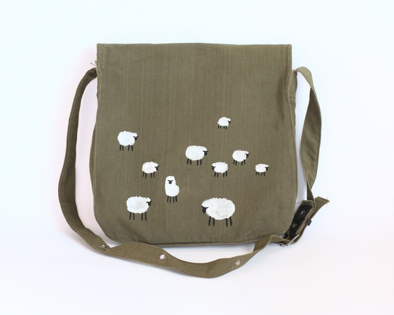 May include: Olive green messenger bag with a flap closure and adjustable strap. The bag features a hand-painted design of white sheep with black legs. The bag is made of a durable fabric and has a snap closure.