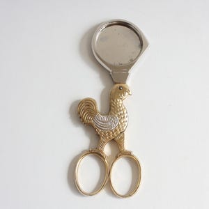 May include: A metal grape scissor set featuring a gold-toned rooster figure with textured feather detailing and silver-toned wings. The handles are formed by two oval gold-toned loops, and the top section includes a circular silver-toned blade.