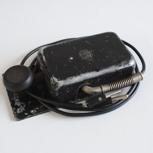 Può includere: Elettrodomestico nero vintage con corpo rettangolare, manico rotondo e manico in metallo a spirale. L'apparecchio ha un cavo di alimentazione e una spina. La superficie mostra segni di usura.