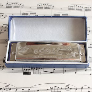 Könnte beinhalten: Ein silbernes Aria Folk Blues Mundharmonika in einer blauen Box. Die Mundharmonika hat eingravierte florale Designs und den Text "The Greatest Little Melody Maker". Die Box ist geöffnet und zeigt ein weißes Innere.