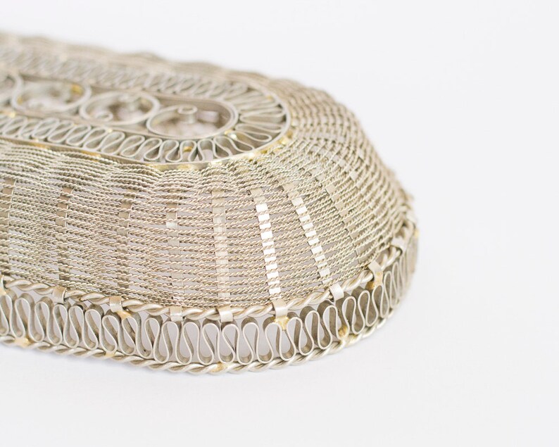 May include: A silver oval-shaped wire basket with a decorative woven pattern. The basket has a scalloped edge with a repeating S-shaped design.