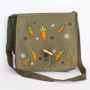 May include: A green canvas messenger bag with a wooden toggle closure. The bag is decorated with hand-painted orange carrots with green tops.