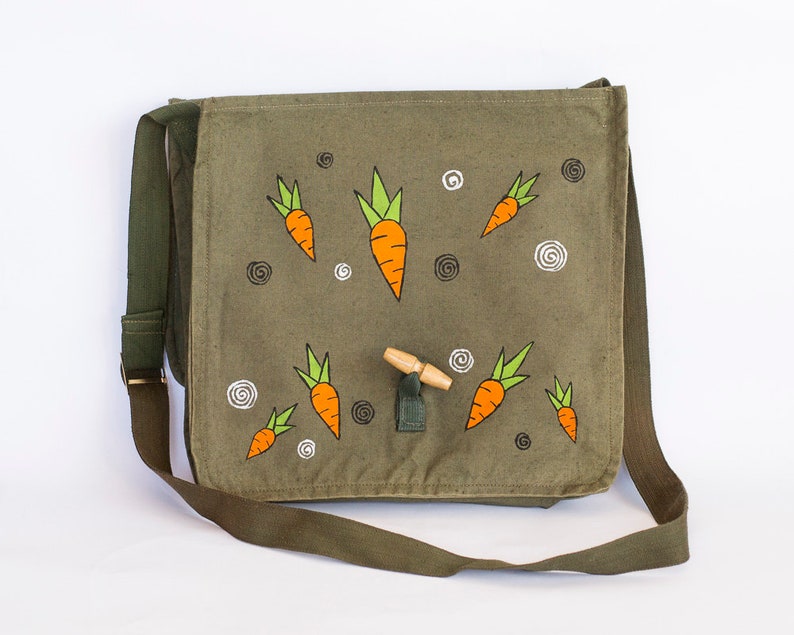 May include: A green canvas messenger bag with a wooden toggle closure. The bag is decorated with hand-painted orange carrots with green tops.