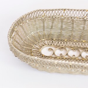 May include: A silver oval-shaped wire basket with a decorative woven pattern. The basket has a detailed border with a repeating design of loops.