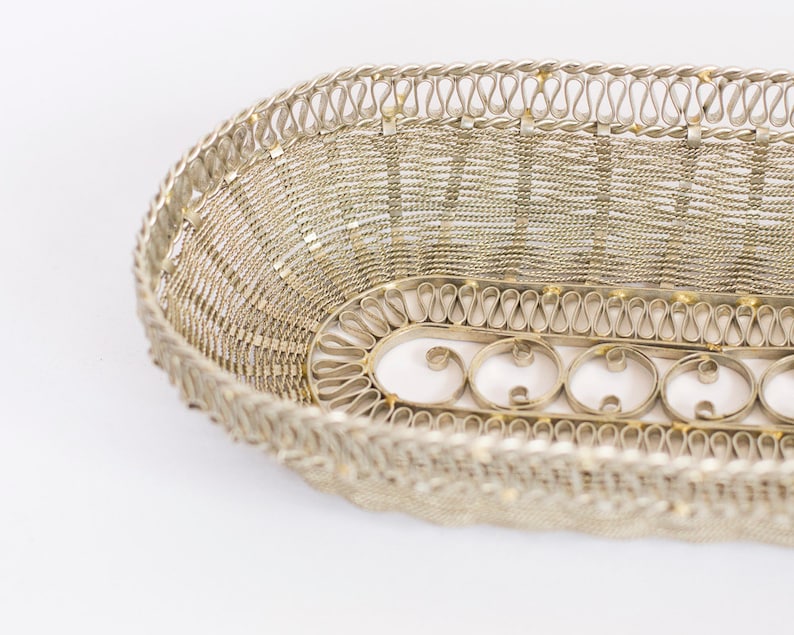May include: A silver oval-shaped wire basket with a decorative woven pattern. The basket has a detailed border with a repeating design of loops.