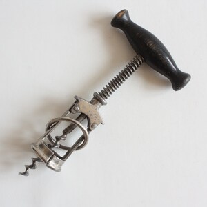 May include: A vintage-style corkscrew featuring a dark brown wooden T-shaped handle, a central metal screw mechanism, and a silver-toned metal frame with a spiral worm. The tool has a mechanical design with a lever-action cage for wine bottles.