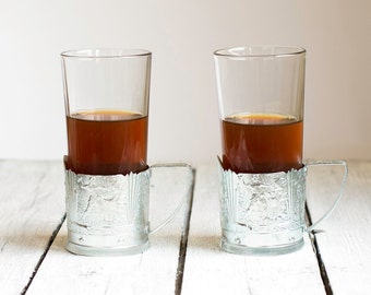May include: Two clear glass cups filled with brown liquid, each sitting in a silver cup holder with an ornate design.