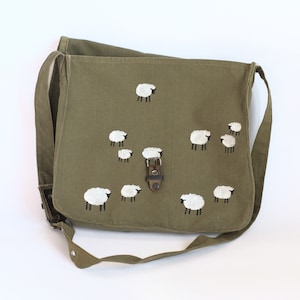 May include: Olive green messenger bag with a flap closure and adjustable shoulder strap. The bag features a hand-painted design of white sheep with black legs. The bag has a metal buckle closure and is made of a durable canvas material.