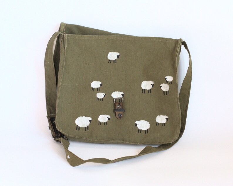 May include: Olive green messenger bag with a flap closure and adjustable shoulder strap. The bag features a hand-painted design of white sheep with black legs. The bag has a metal buckle closure and is made of a durable canvas material.