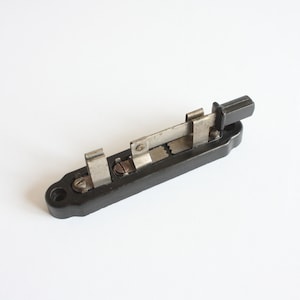 May include: A vintage electrical component featuring a long, rectangular black base with two mounting holes at the ends. It includes a sliding metal mechanism with a black plastic knob, silver-colored metal contact clips, and small screws.
