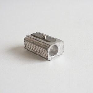 May include: A small, silver pencil sharpener. The sharpener has a rectangular body with ridged sides and a hole for the pencil. A small blade is visible on the top. The sharpener is made of metal and sits on a white surface.