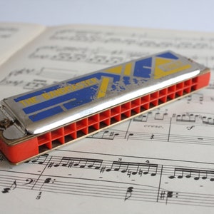 May include: A red and silver harmonica with the words "The Bandmaster" printed on the top. The harmonica is lying on a sheet of music.