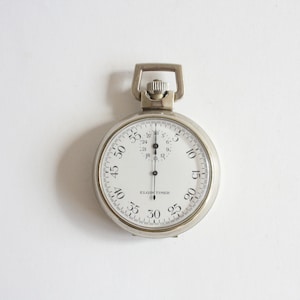 May include: A silver-toned metal stopwatch with a white face, black numerals, and a winding crown at the top. The dial features the text ELGIN TIMER, a large central second hand, and a smaller sub-dial at the top with black markings and numerals.