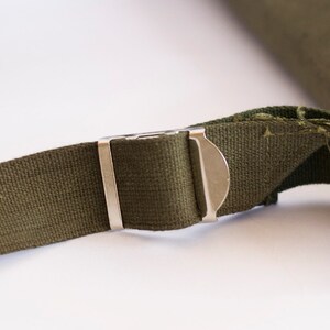 May include: Close-up of a green canvas strap with a silver metal buckle. The strap is frayed at the end.