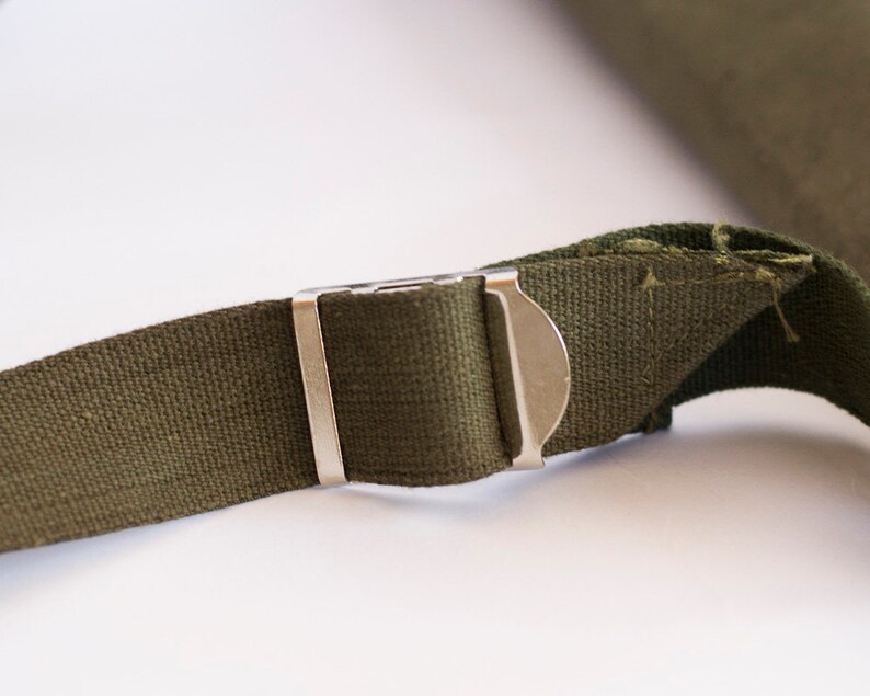 May include: Close-up of a green canvas strap with a silver metal buckle. The strap is frayed at the end.
