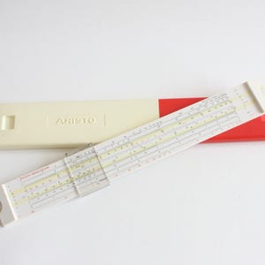 May include: A vintage slide rule with a white and red case. The slide rule has white, black, yellow, and red markings. The case is rectangular, with the word "ARISTO" printed on the top. A clear plastic cursor is on the slide rule.