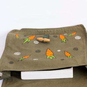 May include: A green canvas bag with a flap closure and a strap. The bag is decorated with hand-painted orange carrots and white swirls. A wooden toggle is visible on the flap.