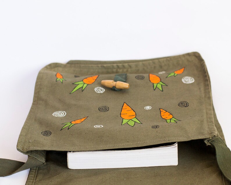 May include: A green canvas bag with a flap closure and a strap. The bag is decorated with hand-painted orange carrots and white swirls. A wooden toggle is visible on the flap.