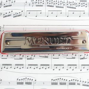 Pode incluir: Uma gaita de boca prateada com uma borda vermelha, repousando sobre partituras. A palavra "MENUET" está gravada na gaita, com "HARMONICA" abaixo. O fundo é preenchido com notas musicais.