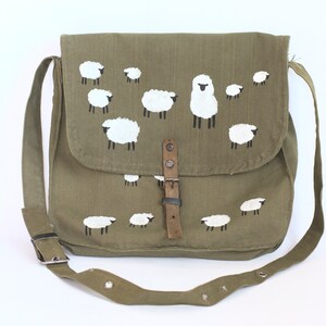 May include: Olive green messenger bag with a flap closure and adjustable strap. The bag features a hand-painted design of white sheep with black legs. The bag has a leather buckle closure and a metal buckle on the strap.