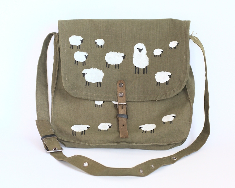 May include: Olive green messenger bag with a flap closure and adjustable strap. The bag features a hand-painted design of white sheep with black legs. The bag has a leather buckle closure and a metal buckle on the strap.
