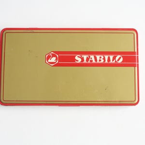 Op de afbeelding: Een rode en gouden metalen doos met het woord "STABILO" in wit op een rode band. Een zwanenlogo bevindt zich in de linkerbovenhoek.