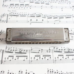 Pode incluir: Uma gaita vintage com o texto "Zigeunervirtuos Wiener Oktavstimmung" em letras prateadas. A gaita repousa sobre uma folha de notas musicais. O instrumento tem formato retangular com corpo castanho escuro e tampa de metal prateado.