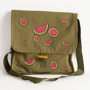 Könnte beinhalten: Eine grüne Canvas-Messenger-Tasche mit einem braunen Lederriemen und einer Holzschnalle. Die Tasche ist mit handgemalten Wassermelonenstücken verziert.