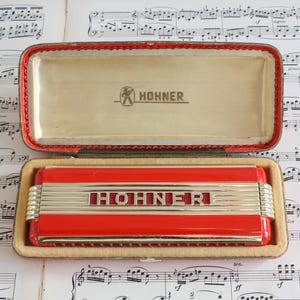 May include: A red and silver Hohner harmonica in an open case. The harmonica has the word "Hohner" in red letters. The case is red with a gold interior and the Hohner logo. The case is open on a background of sheet music.