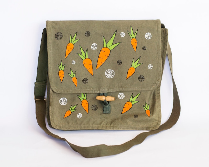 May include: A green canvas messenger bag with a brown leather strap and a wooden toggle closure. The bag is decorated with hand-painted orange carrots with green tops.