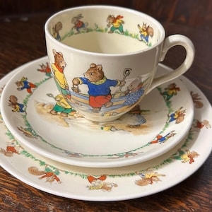 Puede incluir: Un juego de té de cerámica de color crema que consta de una taza, un platillo y un plato. Cada pieza presenta ilustraciones coloridas de animales antropomórficos, incluidos osos y ardillas, realizando actividades al aire libre en la playa.