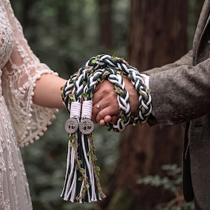 Może przedstawiać: Sznur do ceremonii handfasting z plecionymi białymi i ciemnozielonymi pasmami tkaniny, przeplatanymi małymi zielonymi listkami i sznurkiem. Sznur zakończony jest dwoma okrągłymi metalowymi zawieszkami z motywem drzewa oraz długimi chwostami.