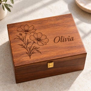 May include: A rectangular wooden box with a hinged lid featuring a laser-engraved floral design of two cosmos flowers and the name Olivia in script font. The box has a smooth, dark brown finish and a small, centered gold-tone metal latch.