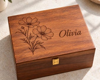 May include: A rectangular wooden box with a hinged lid featuring a laser-engraved floral design of two cosmos flowers and the name Olivia in script font. The box has a smooth, dark brown finish and a small, centred gold-tone metal latch.