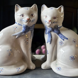 Könnte beinhalten: Zwei weiße Keramik-Katzenfiguren, die nebeneinander sitzen. Jede Katze hat ein handbemaltes blaues Band um den Hals und ein wiederkehrendes Muster aus kleinen blauen und orangefarbenen blumenartigen Motiven auf Körper, Ohren und Stirn.