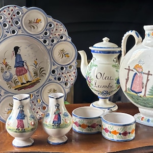 May include: A collection of white ceramic tableware featuring hand-painted folk art designs in blue, red, yellow, and green. The set includes a large pierced decorative plate, two small pitchers, a lidded vessel labelled Oleu Lumbr, and a double bowl.