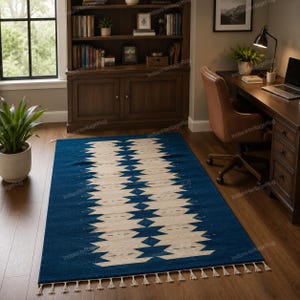May include: A rectangular area rug featuring a deep blue background with a central cream-colored geometric pattern consisting of repeating diamond and zigzag shapes. The rug is finished with short, cream-colored fringe along the short edges.