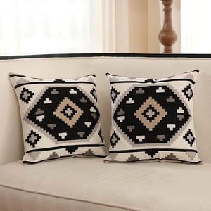 May include: Two square throw pillows featuring a cream-colored base with a prominent black, gray, and tan geometric diamond pattern. The design consists of stacked, angular shapes and horizontal lines that create a symmetrical, repeating motif.