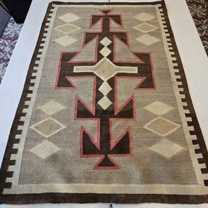 Peut inclure: Un tapis tissé rectangulaire présentant un motif géométrique central en brun foncé et rose sourd sur un fond gris clair. Le design comprend une série de losanges empilés et de formes en flèche, encadrés par une bordure brun foncé à motifs.