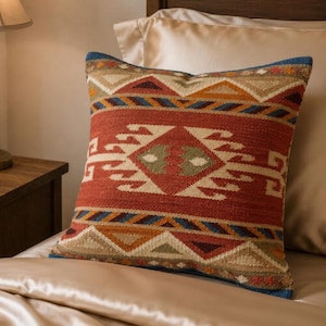 May include: A square throw pillow featuring a woven textile pattern in shades of rust red, tan, olive green, and blue. The design consists of horizontal bands with geometric shapes, including triangles and a central diamond motif, with blue piping.