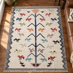 Peut inclure: Un tapis rectangulaire au fond crème orné d'un motif central d'arbre bleu foncé avec des figures d'oiseaux orange, rouges, bleues, vertes et noires disposées symétriquement. Le tapis présente une bordure bleue dentelée et des franges.