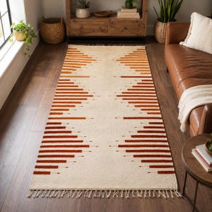 Peut inclure: Un tapis rectangulaire présentant une base de couleur crème avec un motif géométrique symétrique composé de rayures horizontales orange brûlé. Le tapis possède un poil texturé et est fini avec des franges nouées aux extrémités.