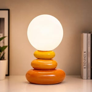 Könnte beinhalten: Eine Schreibtischlampe mit einem weißen, kugelförmigen Glasschirm auf einem Sockel aus drei gestapelten, glatten, abgerundeten Steinen in abgestuften Gelb- und Orangetönen. Der Text CREATIVE DESK LAMP STEAMED WIND STACK STONE ist sichtbar.