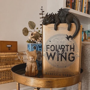 May include: A hardback book titled Fourth Wing with a gold cover, featuring a black dragon figurine perched on top. Beside the book sits a small, light brown carved wooden cat figurine and a pair of clear-framed spectacles on a gold metal table.