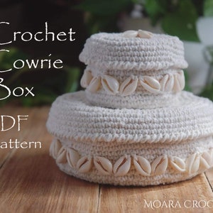 May include: A white crocheted box with a decorative trim of white cowrie shells. The box is stacked on top of another similar box. The text "Crochet Cowrie Box PDF Pattern" is visible in the image.