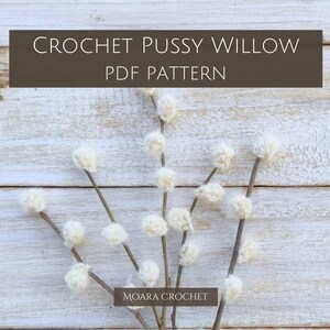May include: Crochet Pussy Willow PDF Pattern by Moara Crochet. A white crocheted pussy willow branch with multiple fluffy white balls on a whitewashed wood background.