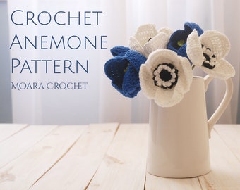 May include: A bouquet of crocheted white and blue anemones in a white pitcher on a wooden table. The text "CROCHET ANEMONE PATTERN MOARA CROCHET" is visible in the image.
