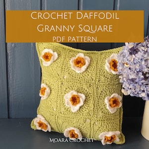 May include: A green crochet granny square pillow with white and orange daffodil flowers. The pillow is on a wooden surface with a blue wall in the background. The text "CROCHET DAFFODIL GRANNY SQUARE PDF PATTERN" is on a brown rectangle above the pillow. The text "MOARA CROCHET" is below the pillow.