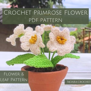 May include: Crochet Primrose Flower PDF Pattern. A close-up of a crocheted primrose flower in a terracotta pot with green leaves. The flower is white with a yellow center. The text "CROCHET PRIMROSE FLOWER PDF PATTERN" is at the top of the image. The text "FLOWER & LEAF PATTERN" is at the bottom left of the image. The text "MOARA CROCHET" is at the bottom right of the image.